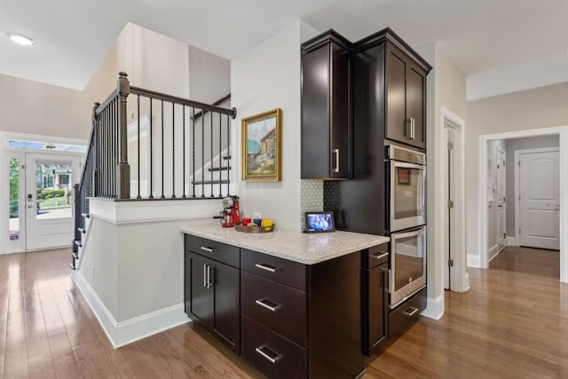 kitchen featuring stainless steel double oven, wood finished floors, light stone counters, baseboards, and dark brown cabinets
