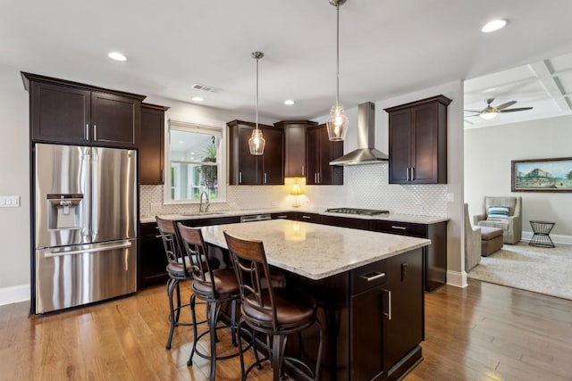 kitchen with stainless steel appliances, wall chimney range hood, dark brown cabinets, light stone countertops, and a sink