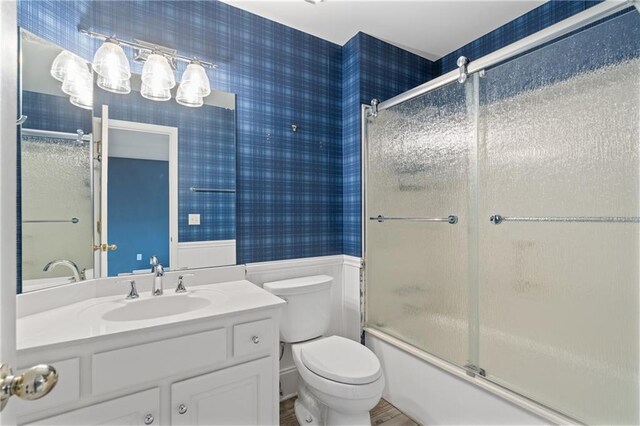 full bathroom with enclosed tub / shower combo, toilet, vanity, wainscoting, and wallpapered walls