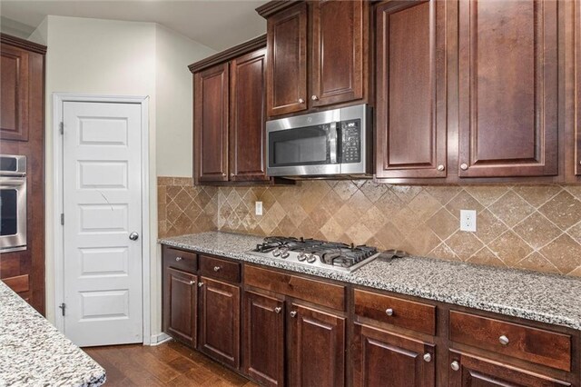 kitchen featuring light stone countertops, decorative backsplash, dark brown cabinets, stainless steel appliances, and dark wood-type flooring