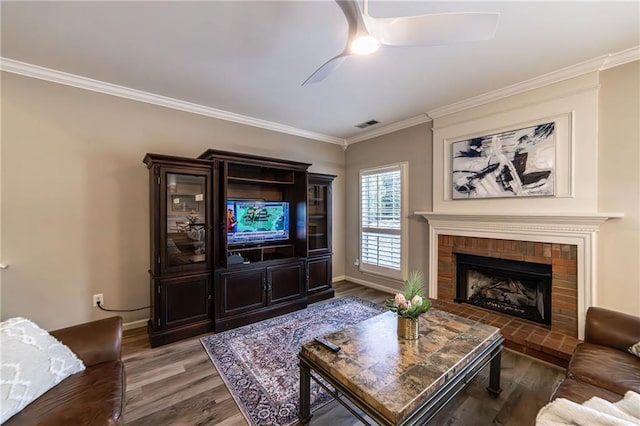 living area featuring baseboards, ceiling fan, wood finished floors, crown molding, and a brick fireplace