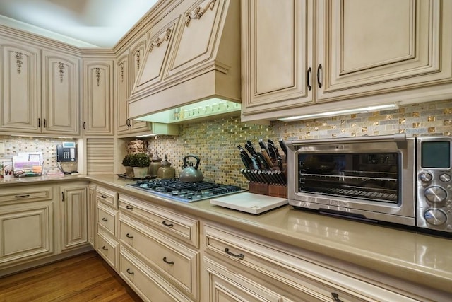 kitchen with cream cabinets, light wood-type flooring, backsplash, premium range hood, and stainless steel gas stovetop