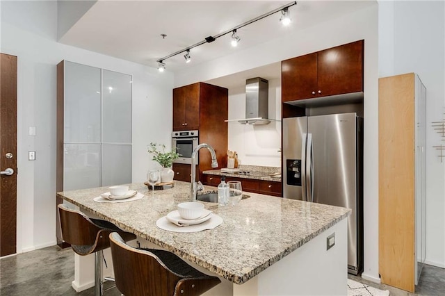 kitchen featuring wall chimney range hood, a sink, an island with sink, stainless steel appliances, and baseboards