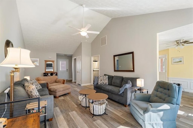 living area with ceiling fan, high vaulted ceiling, a wainscoted wall, wood finished floors, and visible vents