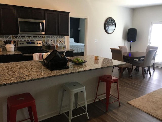 kitchen featuring dark wood-type flooring, dark stone countertops, appliances with stainless steel finishes, tasteful backsplash, and a kitchen bar