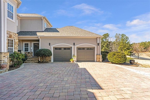 exterior space with a shingled roof, stucco siding, a garage, stone siding, and decorative driveway