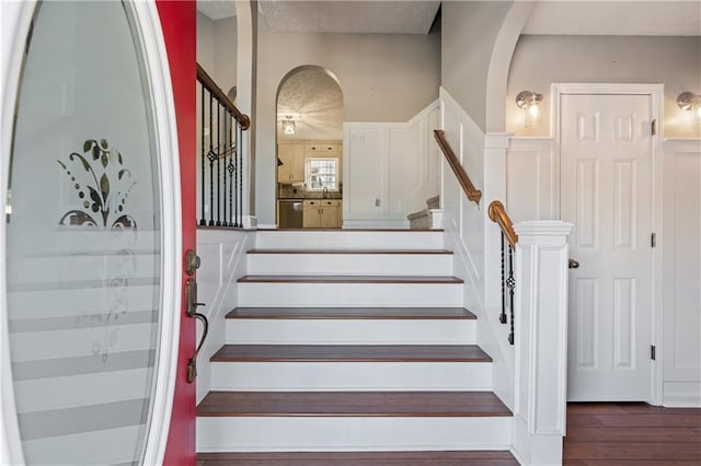 staircase with a wainscoted wall, arched walkways, and wood finished floors