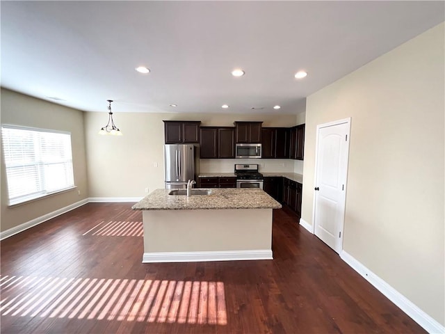 kitchen with stainless steel appliances, a sink, an island with sink, light stone counters, and dark brown cabinetry