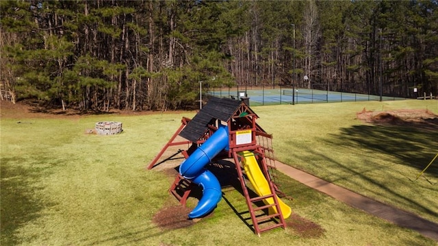 community jungle gym featuring a tennis court, a lawn, a forest view, and fence