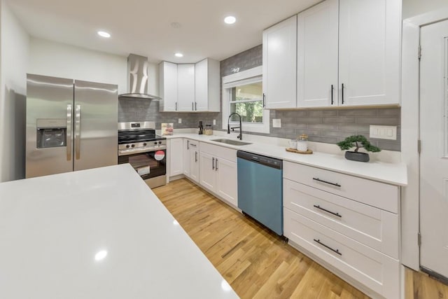 kitchen with sink, white cabinetry, stainless steel appliances, light hardwood / wood-style floors, and wall chimney range hood