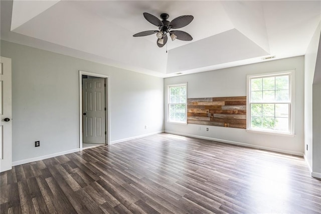 empty room with a raised ceiling, healthy amount of natural light, a ceiling fan, and dark wood-type flooring