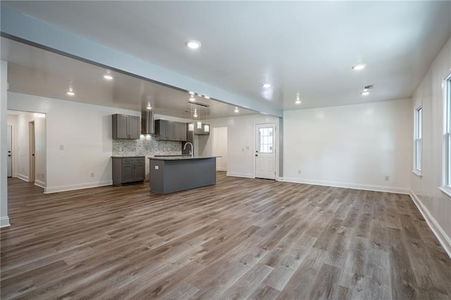 kitchen featuring backsplash, wall chimney exhaust hood, a kitchen island with sink, wood-type flooring, and gray cabinets