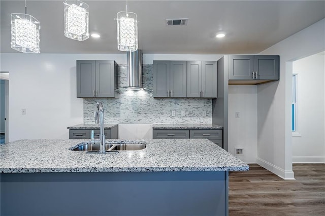 kitchen with light stone counters, sink, wall chimney range hood, pendant lighting, and gray cabinets