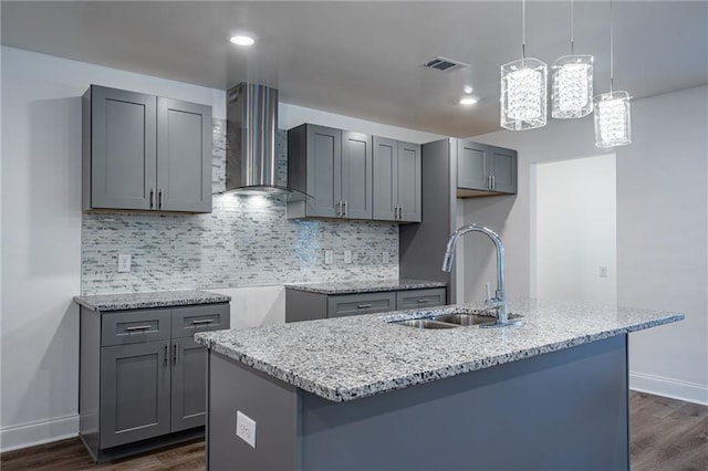 kitchen with light stone countertops, sink, wall chimney range hood, gray cabinets, and a kitchen island with sink