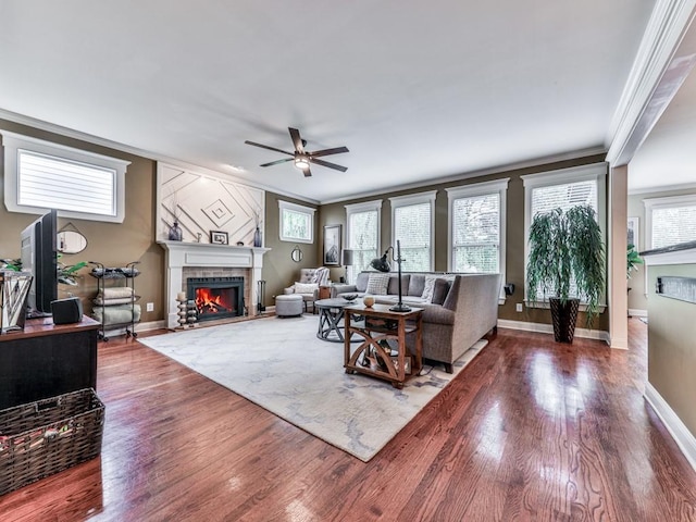 living area featuring a warm lit fireplace, ceiling fan, crown molding, wood finished floors, and baseboards