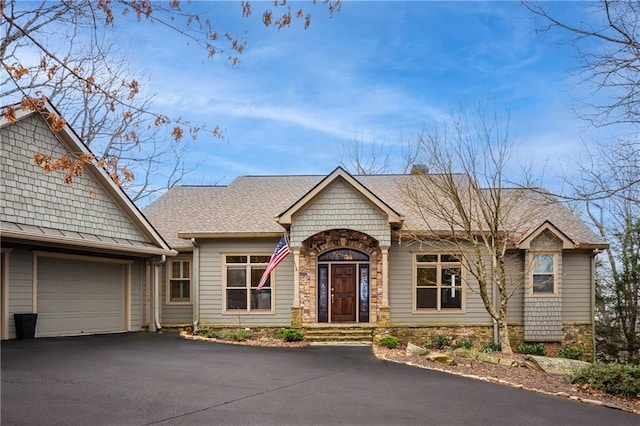 ranch-style home featuring driveway, stone siding, an attached garage, and roof with shingles