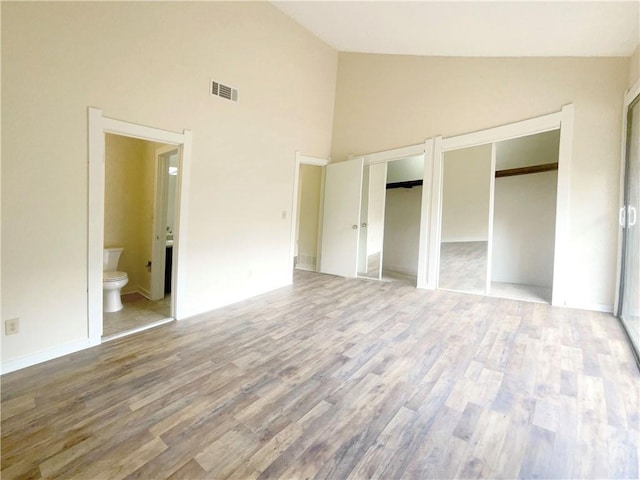 unfurnished bedroom featuring high vaulted ceiling, ensuite bathroom, hardwood / wood-style floors, and two closets