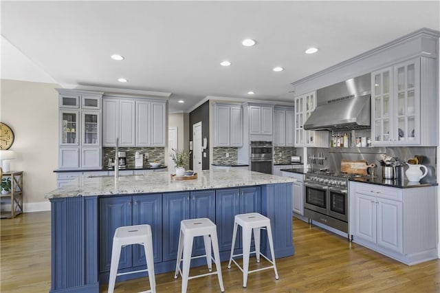 kitchen with a large island with sink, wall chimney range hood, light wood-style flooring, and appliances with stainless steel finishes