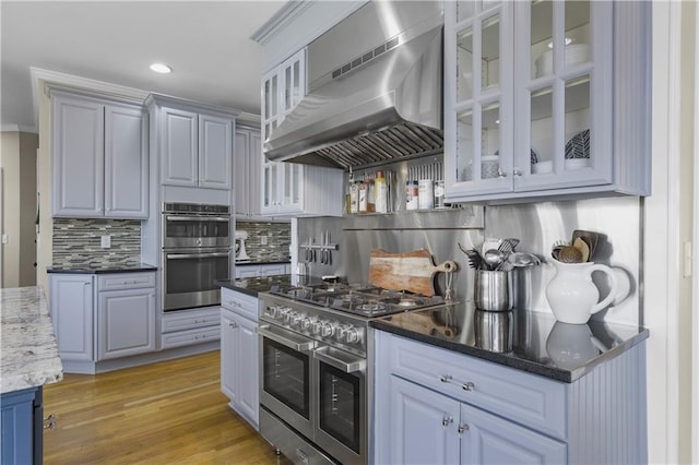 kitchen with dark stone counters, stainless steel appliances, light wood-style floors, wall chimney exhaust hood, and tasteful backsplash