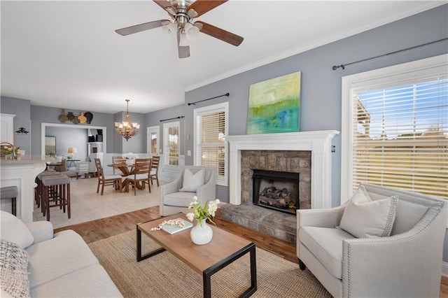 living area featuring light wood-style floors, a stone fireplace, ornamental molding, and ceiling fan with notable chandelier