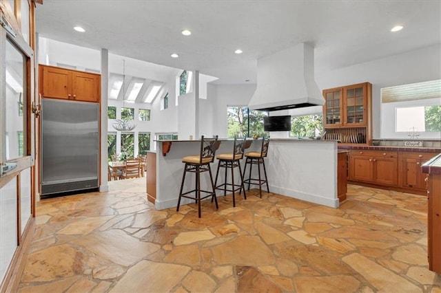 kitchen with a breakfast bar area, stainless steel built in fridge, custom exhaust hood, and a healthy amount of sunlight