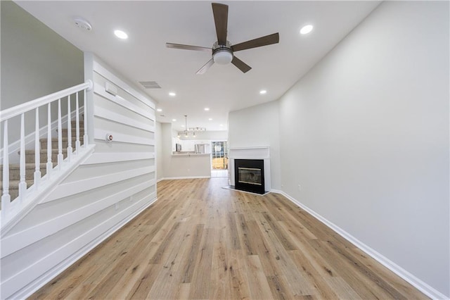 unfurnished living room featuring light wood finished floors, visible vents, a glass covered fireplace, ceiling fan, and stairs