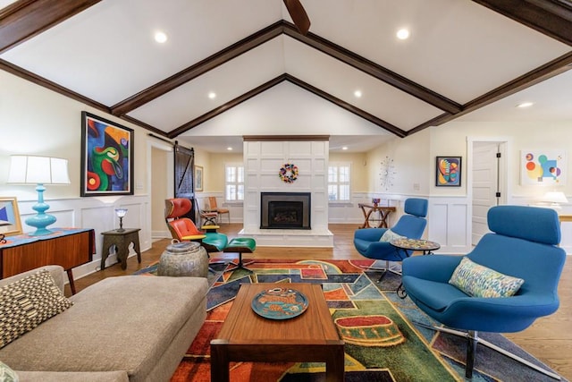 living area with a barn door, a fireplace with raised hearth, a wainscoted wall, wood finished floors, and vaulted ceiling with beams