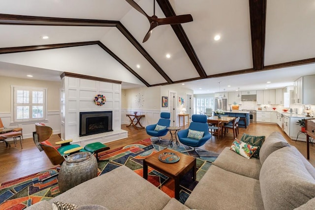 living area with light wood-type flooring, a wainscoted wall, a fireplace with raised hearth, and beamed ceiling