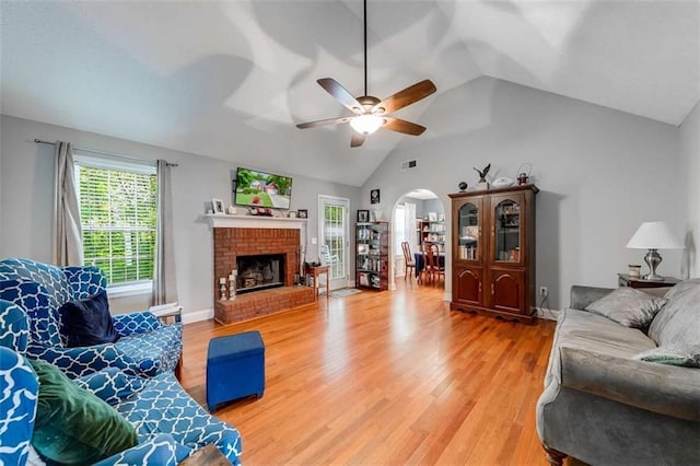 living area with a ceiling fan, a brick fireplace, light wood-style floors, arched walkways, and vaulted ceiling
