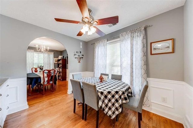dining area with light wood-style floors, arched walkways, wainscoting, and ceiling fan with notable chandelier