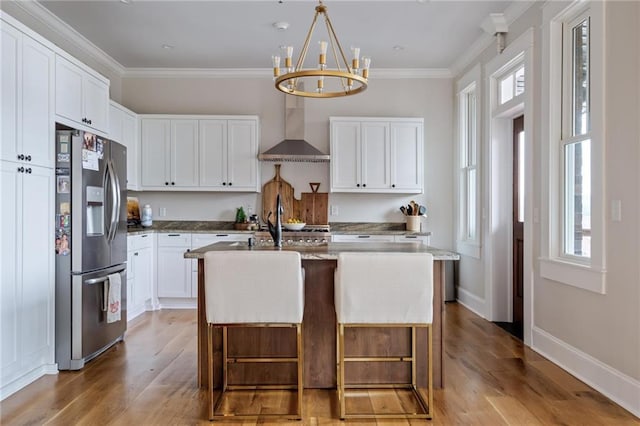 kitchen with stainless steel fridge, a center island with sink, wall chimney exhaust hood, a kitchen bar, and white cabinetry