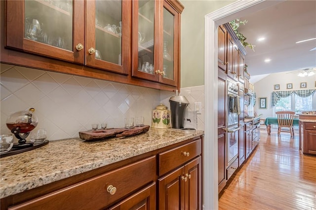 kitchen with light wood finished floors, backsplash, glass insert cabinets, light stone counters, and brown cabinetry