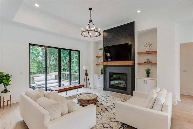 living room featuring a large fireplace, a raised ceiling, light wood-type flooring, a notable chandelier, and built in shelves