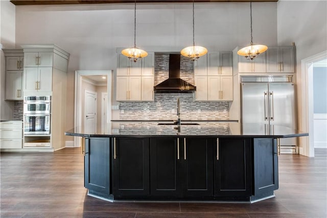 kitchen with wall chimney exhaust hood, a kitchen island with sink, appliances with stainless steel finishes, and dark stone counters