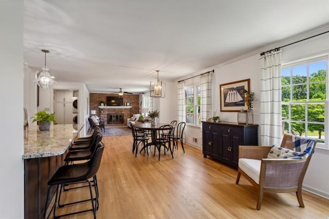 dining space featuring stacked washer and clothes dryer, ceiling fan with notable chandelier, light wood-type flooring, and a brick fireplace