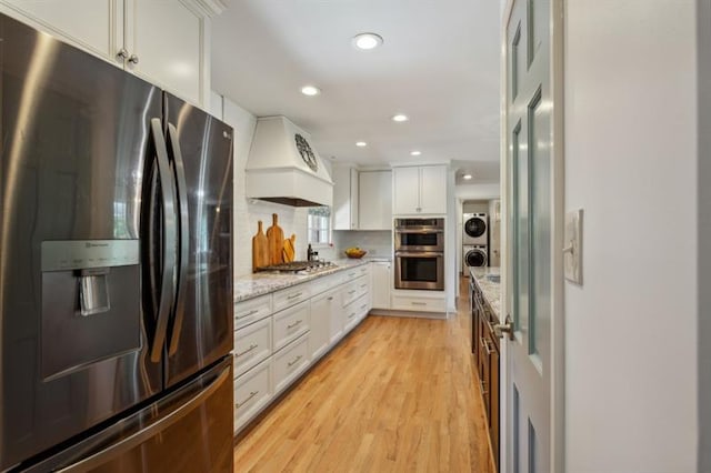 kitchen with white cabinetry, custom exhaust hood, stacked washer and clothes dryer, light stone counters, and stainless steel appliances