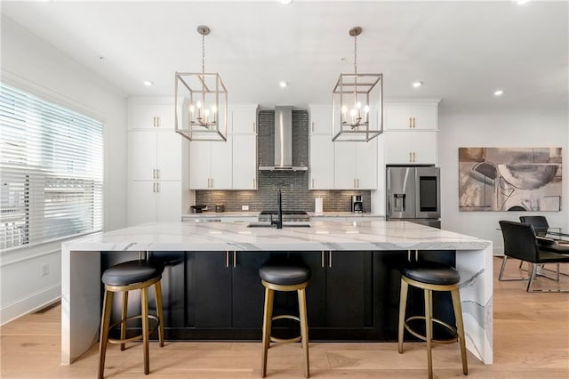 kitchen with stainless steel fridge with ice dispenser, a large island with sink, decorative backsplash, white cabinetry, and wall chimney range hood