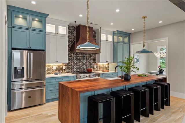 kitchen featuring blue cabinetry, custom range hood, wooden counters, backsplash, and stainless steel fridge