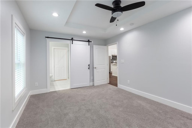 unfurnished bedroom featuring light carpet, ceiling fan, a raised ceiling, a barn door, and connected bathroom