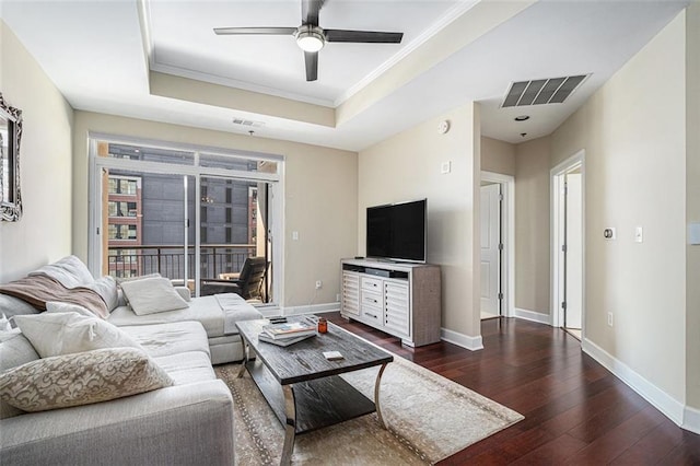 living area with a raised ceiling, dark wood-type flooring, ceiling fan, crown molding, and baseboards