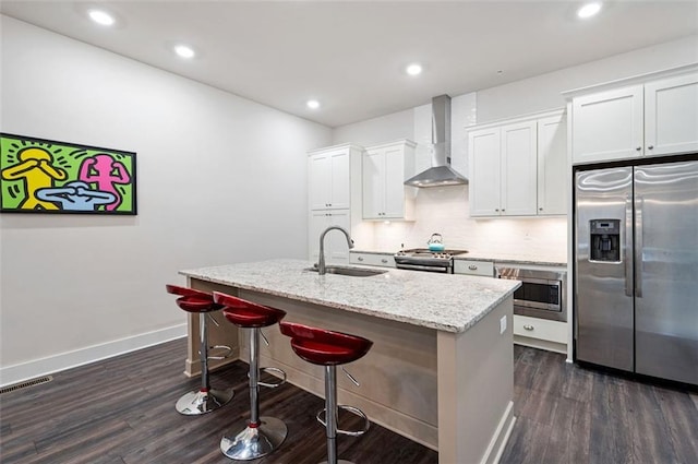 kitchen featuring dark wood-type flooring, sink, an island with sink, stainless steel appliances, and wall chimney range hood