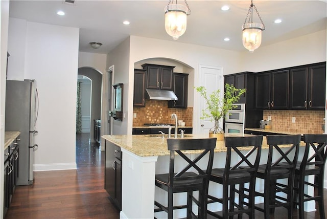 kitchen with stainless steel appliances, an island with sink, light stone counters, and decorative light fixtures