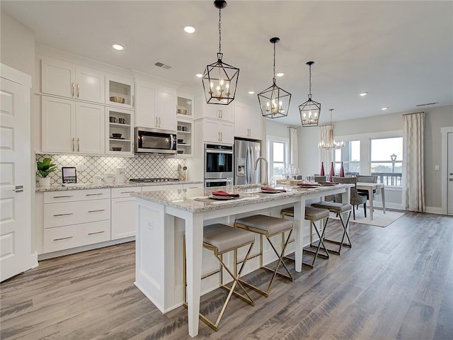 kitchen featuring stainless steel appliances, light stone counters, a center island with sink, light wood-type flooring, and pendant lighting