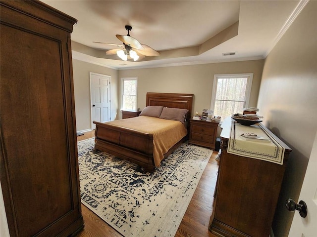 bedroom featuring a tray ceiling, crown molding, dark wood finished floors, visible vents, and a ceiling fan