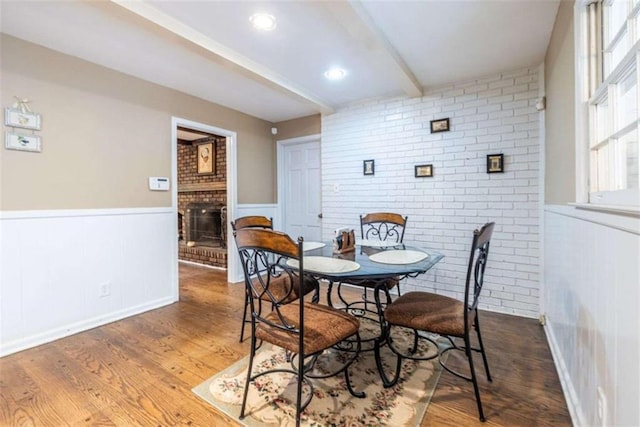 dining space featuring a brick fireplace, wood finished floors, beamed ceiling, wainscoting, and brick wall