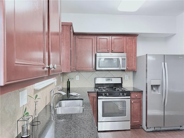 kitchen with appliances with stainless steel finishes, a sink, reddish brown cabinets, dark stone countertops, and tasteful backsplash