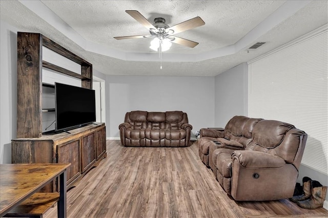 living room with ceiling fan, a textured ceiling, a tray ceiling, and light wood-type flooring