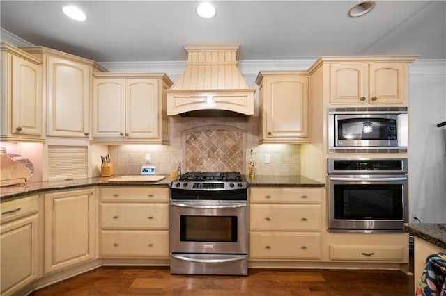 kitchen with dark stone counters, stainless steel appliances, cream cabinetry, premium range hood, and backsplash