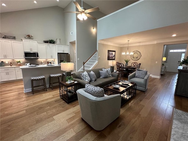 living room with crown molding, ceiling fan with notable chandelier, light wood-style floors, baseboards, and stairs