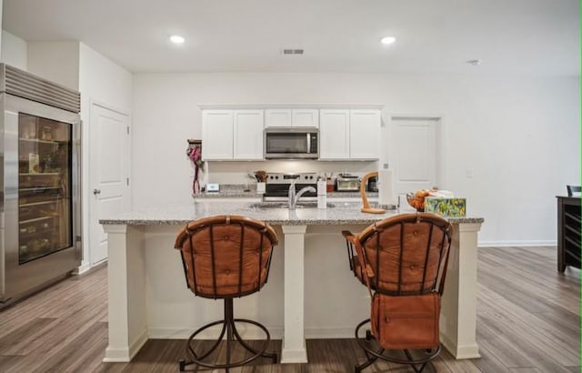 kitchen featuring hardwood / wood-style flooring, light stone counters, appliances with stainless steel finishes, an island with sink, and white cabinets